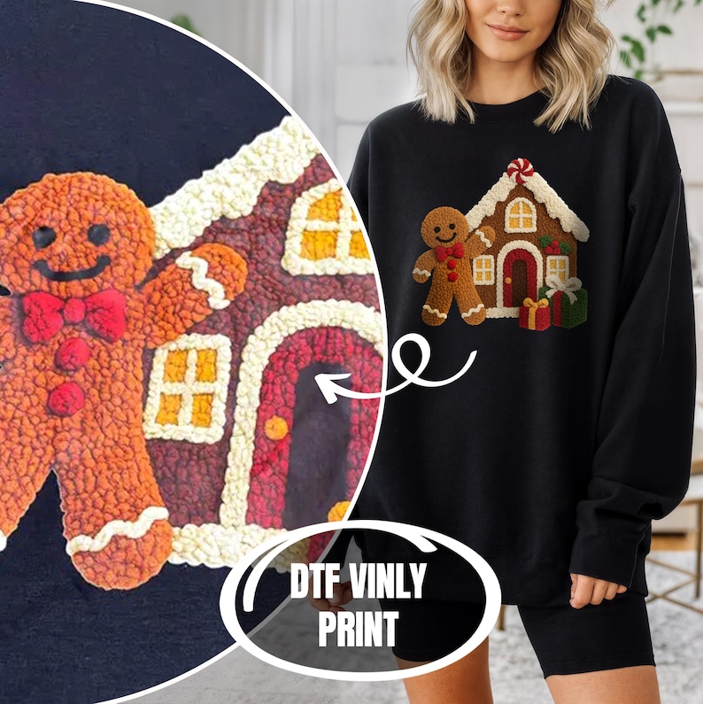 A woman wearing a black sweater with a gingerbread house design printed on it, standing next to a large gingerbread house.