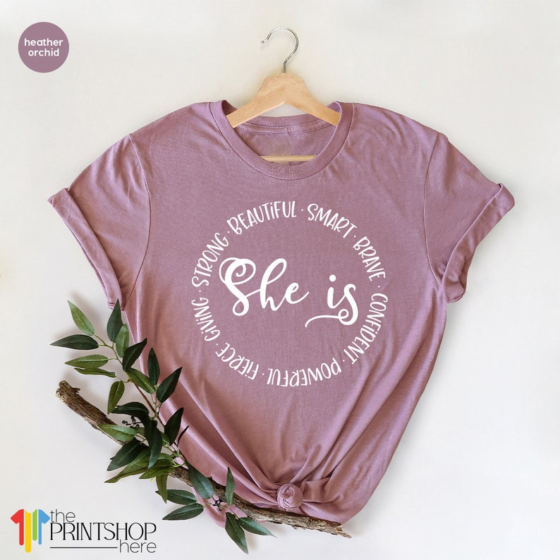 May include: A heather orchid t-shirt with a white circle graphic that says "She is" surrounded by the words "powerful, fierce, giving, strong, beautiful, smart, brave, confident".