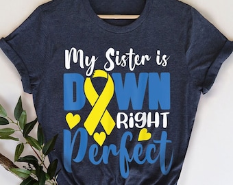 May include: A navy blue t-shirt with the text "My Sister is Down Right Perfect" in white and blue lettering. A yellow ribbon is in the center of the text. The ribbon is a symbol of Down Syndrome awareness.