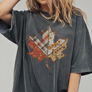 May include: A charcoal gray short-sleeve t-shirt featuring a large maple leaf graphic on the front. The leaf design is divided into sections with various patterns including plaid, floral, and pumpkin prints in shades of orange, brown, and cream.
