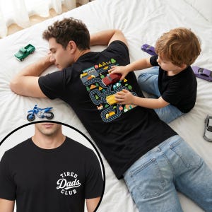 May include: Black t-shirt with a road map design on the back and the words "Tired Dads Club" on the front. A child plays with toy cars on the shirt. The shirt is worn by a person lying on a white bed.