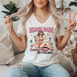 May include: A cream-colored V-neck t-shirt featuring a graphic of a person in a polka-dot swimsuit sitting on a beach towel. The text reads BEACH BABE and Catching Rays, NOT FEELINGS, surrounded by illustrations of palm trees, a sun, and stars.