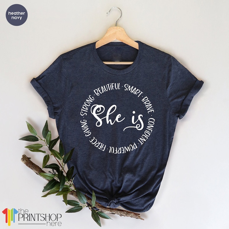 May include: A heather navy blue t-shirt with a white circle graphic that says "She is" with the words "powerful, fierce, giving, strong, beautiful, smart, brave, confident" surrounding the text.