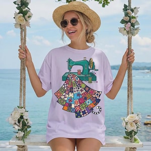 May include: A light purple t-shirt featuring a graphic illustration of a teal sewing machine stitching a colorful, multi-patterned quilt. The design includes small spools of thread and a vibrant patchwork quilt design with various geometric shapes.