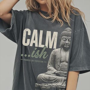 May include: A charcoal gray short-sleeve t-shirt featuring a graphic of a seated Buddha statue. The text reads CALM in large white letters, followed by ...ish in green script, and the phrase PROGRESS, NOT PERFECTION in small white letters.