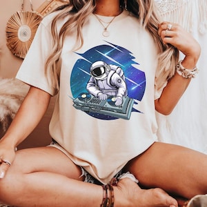 May include: Beige t-shirt featuring a graphic of an astronaut DJing in space. The astronaut is wearing a white spacesuit and is positioned in front of a blue and purple galaxy background with shooting stars. The DJ setup is gray.