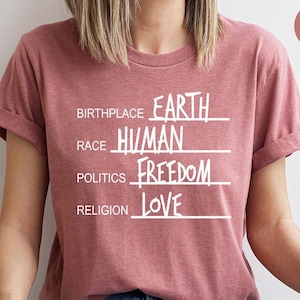 May include: A heather pink t-shirt with white text that reads "BIRTHPLACE EARTH, RACE HUMAN, POLITICS FREEDOM, RELIGION LOVE".