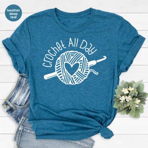 May include: A teal blue t-shirt with the text "Crochet All Day" in white. The design features a ball of yarn with a heart in the center and a crochet hook.