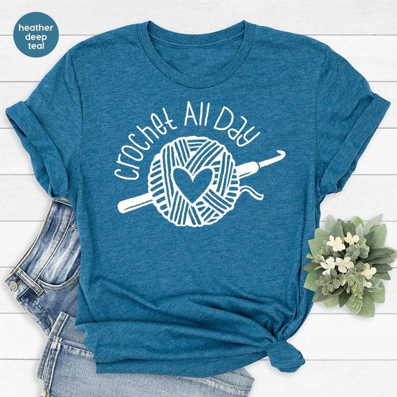 May include: A teal blue t-shirt with the text "Crochet All Day" in white. The design features a ball of yarn with a heart in the center and a crochet hook.