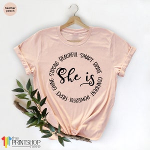 May include: A peach colored t-shirt with a black circle graphic that says "She is" surrounded by the words "Strong, Beautiful, Smart, Brave, Confident, Powerful, Fierce, Giving".
