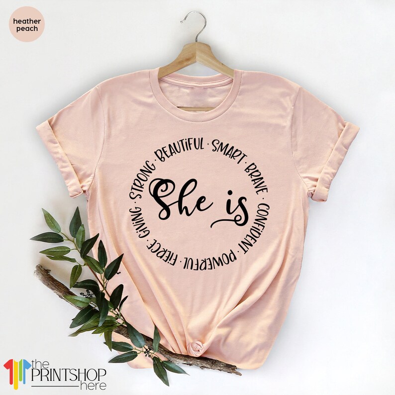 May include: A peach colored t-shirt with a black circle graphic that says "She is" surrounded by the words "Strong, Beautiful, Smart, Brave, Confident, Powerful, Fierce, Giving".