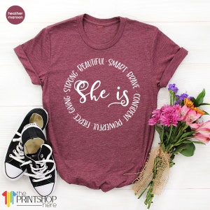 May include: Heather maroon t-shirt with a white circle graphic that says "She is" surrounded by the words "powerful, fierce, giving, strong, beautiful, smart, brave, confident".