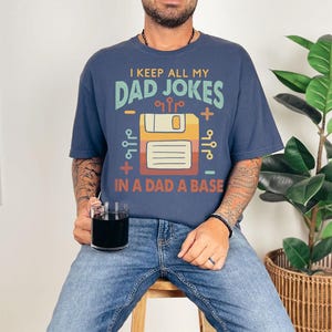 May include: Navy blue t-shirt with the text "I KEEP ALL MY DAD JOKES IN A DAD A BASE" and a retro floppy disk graphic. The shirt has a comfortable fit and a crew neck.