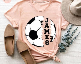 May include: A peach colored t-shirt with a black and white soccer ball graphic. The soccer ball is cut in half and the name "JAMES" is written vertically on the right side of the graphic.