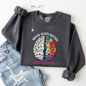 May include: Dark gray sweatshirt with the text "MENTAL HEALTH MATTERS" and a graphic of a brain split into two halves. One half is white, the other is a colorful rainbow design. The hashtag "#ENDTHESTIGMA" is below the brain graphic.