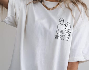 Puede incluir: Camiseta de color crema con un dibujo lineal negro de una pareja abrazándose. La ilustración representa a un hombre y una mujer en un momento tierno. La camiseta tiene un corte holgado y cuello redondo.