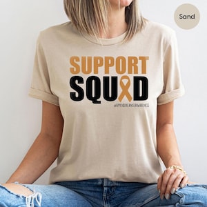 May include: A beige t-shirt with the text "SUPPORT SQUAD" in black letters and a brown ribbon. The hashtag #APPENDICANCERAWARENESS is printed below the text.