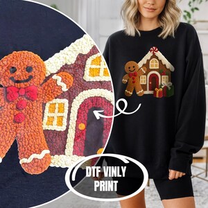 A woman wearing a black sweater with a gingerbread house design printed on it, standing next to a large gingerbread house.