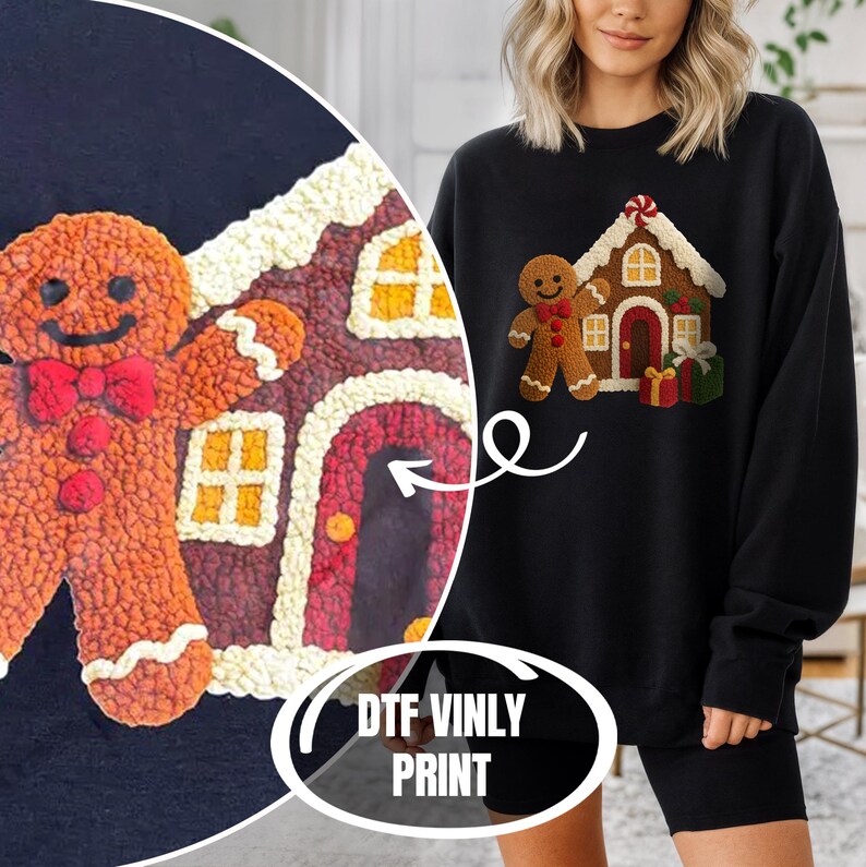 A woman wearing a black sweater with a gingerbread house design printed on it, standing next to a large gingerbread house.