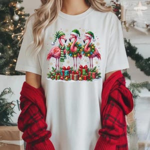 May include: A white short-sleeve t-shirt featuring a graphic of three pink flamingos wearing Christmas hats. The flamingos are decorated with a green scarf and wreaths with colorful lights, standing above a pile of wrapped gift boxes.