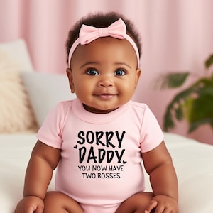 Może przedstawiać: Uśmiechnięte dziecko w jasnoróżowym body i pasującą opaską z kokardą. Body ma czarny napis "SORRY DADDY. YOU NOW HAVE TWO BOSSES". Tło jest różowe.