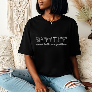 May include: Black t-shirt with white line art of medical symbols and the text "women's health nurse practitioner". The design includes a stethoscope, flowers, and reproductive health symbols. The shirt is a casual, oversized fit.