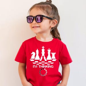 Peut inclure: Un enfant porte des lunettes de soleil noires et un t-shirt rouge avec un motif d'échecs blanc. Le motif comprend des pièces d'échecs, le texte "I'M THINKING" et un cercle de chargement. Le t-shirt est de couleur rouge vif.