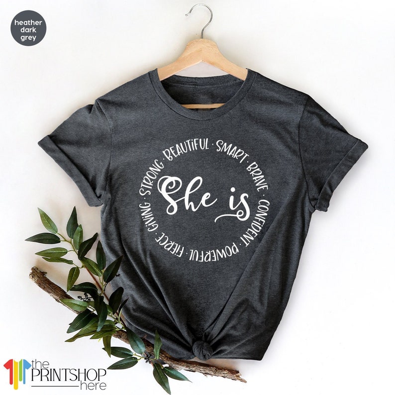 May include: A heather dark grey t-shirt with a white circle graphic that says "She is" surrounded by the words "powerful, fierce, giving, strong, beautiful, smart, brave, confident".