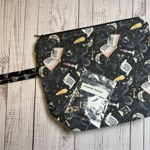Puede incluir: Bolsa negra con cremallera con un estampado negro y dorado de cuervos, llaves, lunas y libros abiertos. Una pequeña bolsa de plástico transparente etiquetada como "Lavender Sachet" está encima de la bolsa.