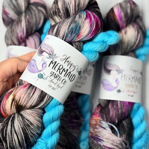 May include: Skeins of hand-dyed yarn with a mermaid label. The yarn is a mix of gray, black, pink, and blue. The label reads "Happy Mermaid Yarn Co." and features a mermaid illustration. The yarn is a vibrant and colorful craft supply.