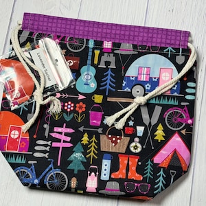 May include: A black drawstring bag with a camping-themed pattern featuring bicycles, tents, and camper vans. The bag has a purple top and a white drawstring. A small package labeled "Lavender Sachet" is attached.
