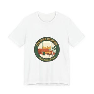 Könnte beinhalten: Weißes T-Shirt mit einem kreisförmigen Aufdruck. Der Aufdruck zeigt den Text "CERTIFIED INDOORSY" und "Couch National Park". Es zeigt eine Wohnzimmerszene mit Sofa, Couchtisch und Pflanzen. Ein Fenster zeigt eine Stadtlandschaft.
