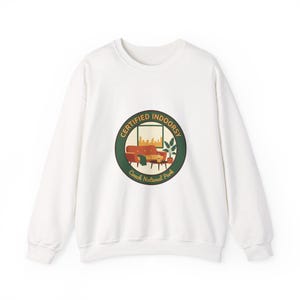 Könnte beinhalten: Weißes Sweatshirt mit einem kreisförmigen Aufdruck mit der Aufschrift "CERTIFIED INDOORSY" um ein Design mit Sofa, Couchtisch und Stadtlandschaft. Der Text "Couch National Park" befindet sich am unteren Rand der Grafik.