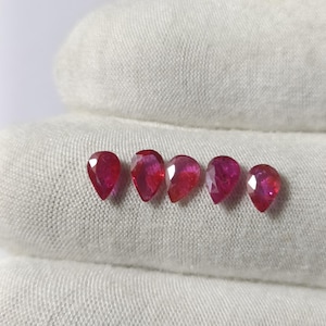 May include: Five pear-shaped, faceted rubies are displayed on a white cloth. The gemstones are a deep, vibrant red colour, with visible internal facets. The gems are arranged in a row, showcasing their polished surfaces and unique shapes.