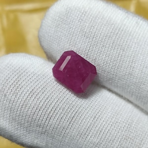 May include: A faceted, rectangular ruby gemstone. The gem is a deep, rich red color and has multiple facets that catch the light. The ruby is held against a white fabric background.