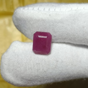 May include: A deep red, rectangular-cut ruby gemstone. The gem is displayed against a white fabric background, highlighting its colour and clarity. The emerald cut features a flat top and beveled edges. The stone is likely to be used in jewellery.