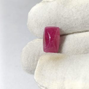 May include: A rectangular, polished ruby gemstone with rounded corners. The gem is a deep, saturated pinkish-red color. The stone is held against a white fabric background.