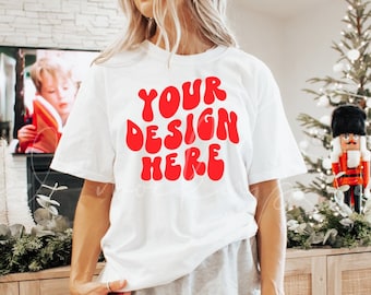 May include: A white t-shirt with the text "YOUR DESIGN HERE" in red, wavy letters. The shirt is being worn by a person standing in front of a wooden cabinet with a woven rattan front.