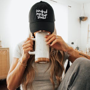 Könnte beinhalten: Eine Person trägt eine schwarze Baseballkappe mit weißem Text, der "kindred mockup studio" lautet, und trinkt aus einer weißen Keramiktasse mit schwarzen Flecken. Die Person trägt ein graues Shirt und graue Jogginghosen.
