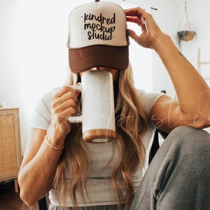 Puede incluir: Una mujer lleva una gorra de camionero marrón y blanca con el texto "kindred mockup studio" en letras negras. Está sosteniendo una taza de cerámica blanca con detalles marrones.