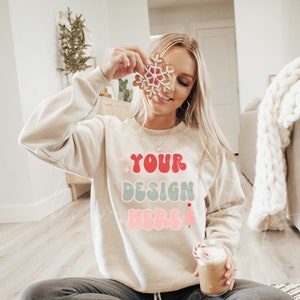 K&ouml;nnte beinhalten: Eine Frau tr&auml;gt ein cremefarbenes Sweatshirt mit dem Text "YOUR DESIGN HERE" in Rot, Rosa und Blau. Sie h&auml;lt ein Lebkuchen-Schneeflocken-Kekse vor ihrem Gesicht und sitzt auf einem schwarz-wei&szlig;en Teppich.