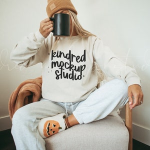 May include: A woman wearing a beige sweatshirt with the text "kindred mockup studio" in black lettering. She is sitting in a chair with a blanket on her lap and is holding a black mug.