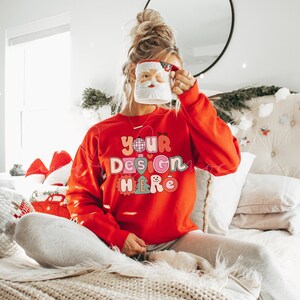 May include: A woman wearing a red sweatshirt with the text "YOUR DESIGN HERE" in colorful letters. She is holding a white mug with a Santa Claus face on it. The woman is sitting on a bed with white bedding and a white headboard. There is a small Christmas tree in the background.