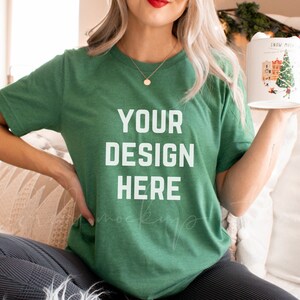 May include: A woman wearing a green t-shirt with the text "YOUR DESIGN HERE" on it. She is sitting on a white blanket and holding a mug with a Christmas design.