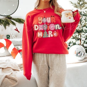 May include: A red sweatshirt with the text "your design here" in colourful letters with holiday-themed graphics. The sweatshirt is being worn by a person in a casual setting.
