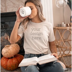Könnte beinhalten: Eine Frau trägt ein beiges T-Shirt mit dem Aufdruck "your design here". Sie sitzt auf einem Teppich mit einem Buch auf dem Schoß und einem Kürbis neben sich.
