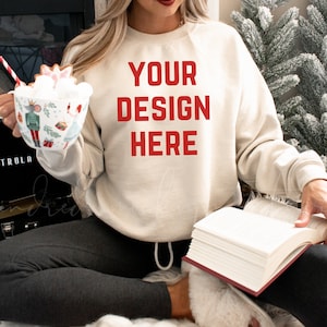 May include: A woman wearing a beige sweatshirt with the text "YOUR DESIGN HERE" in red. She is holding a mug with a winter scene and a book.