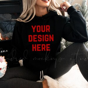 May include: A black hooded sweatshirt with the text "YOUR DESIGN HERE" in red lettering. The sweatshirt is being worn by a person sitting in front of a fireplace.