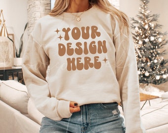 Puede incluir: Una sudadera de color crema con el texto "YOUR DESIGN HERE" en letras marrones con una fuente retro. La sudadera la lleva puesta una persona con el pelo largo y castaño. La persona está de pie delante de un árbol de Navidad.