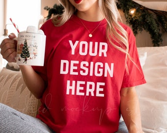 Könnte beinhalten: Ein rotes T-Shirt mit dem Aufdruck "YOUR DESIGN HERE" auf der Vorderseite. Das Shirt wird von einer Person getragen, die auf einem Bett mit einer weißen Decke und Kissen sitzt.